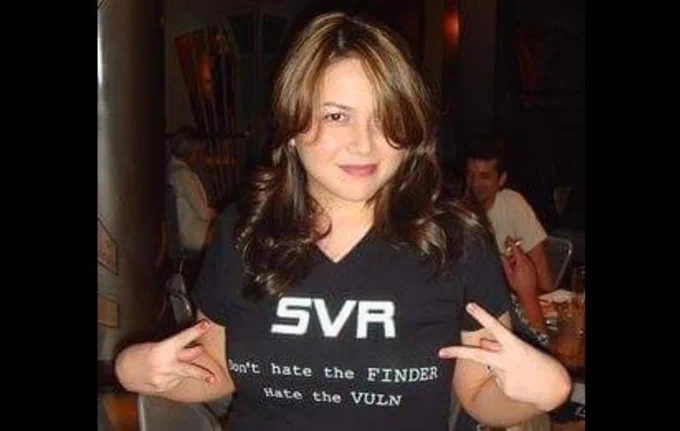 Katie with brown hair holding up two hands with the V peace sign in America and meaning something else in England, wearing a black V neck tshirt with white letters that say:
SVR
Don’t hate the finder 
Hate the vuln