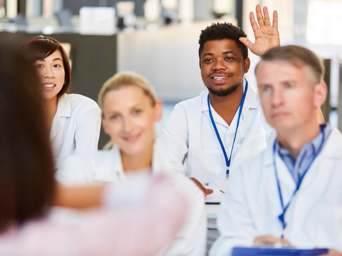 A group of people in lab coats in a classroom. One individual is raising a hand while others are attentively looking forward. 