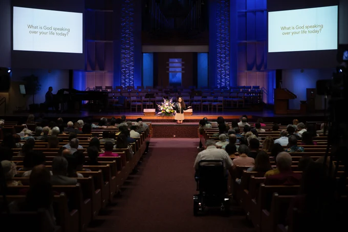 A large audience is seated in a church auditorium facing a stage. Two screens display the question "What is God speaking over your life today?" A person is speaking at the podium, with a floral arrangement nearby. The room is dimly lit with a focus on the stage area.