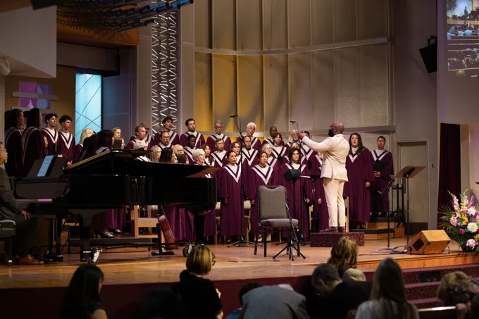 A choir dressed in burgundy robes is singing on a church stage. A conductor in light-colored attire stands in front, leading them. A grand piano is on the left, and a floral arrangement is on the right. A cross design is visible in the background. Audience members are seated in the foreground.