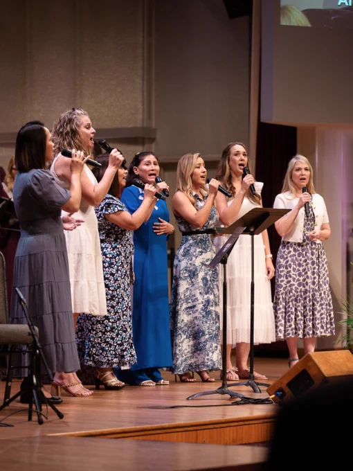 A group of woman are standing on a stage, singing into microphones. They are wearing dresses and are positioned behind a music stand.