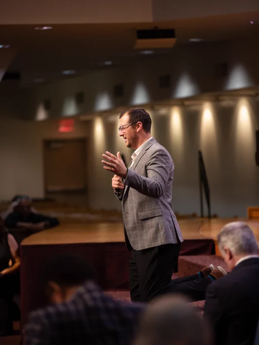 A person in a grey blazer and glasses is speaking into a microphone on a church stage. He is kneeling while praying with an audience seated in front.