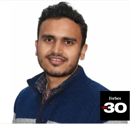 Portrait of Puneet Singhal wearing a navy blue sweater over a checked shirt, shown against a white background. A Forbes Under 30 logo appears in the bottom-right corner.