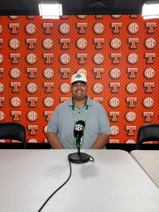 The image shows a person sitting behind a white table in a press conference setting. The background consists of an orange wall with a repeating pattern of "S-E-C" logos and the letter "T," both in white circles. The person is wearing a white cap with green text, glasses, and a light-colored button-up shirt. In front of them on the table is a microphone attached to a stand with the S-E-C logo. The setting is brightly lit with two square lights visible at the top of the image.