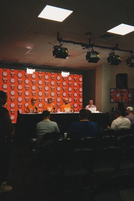 The image shows a press conference setting with a panel of four people sitting behind a long black table. Three of them are wearing matching orange shirts and baseball caps with the text "Vols" on the shirts. The backdrop consists of a pattern featuring an orange logo "T" and "S-E-C" on a red background. Overhead, there are several lighting fixtures illuminating the panel. An audience is seated in front of the panel, facing them. To the right, a person, likely a journalist, is sitting at a separate table with a laptop. A large speaker stands in the corner of the room, with a television screen displaying the panel members.
