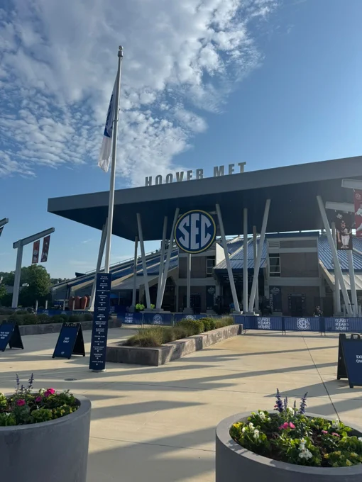 The image depicts the entrance of Hoover Metropolitan Stadium, a structure adorned with a sign reading "HOOVER MET" on top. The foreground features a wide walkway lined with large concrete planters holding colorful flowers. In the middle ground, a tall flagpole with a white flag stands prominently. Below the stadium's roofline is a round S-E-C logo. The stadium itself is composed of modern architectural elements, including a slanted metal roof supported by angled white beams. The sky above is mostly clear with scattered clouds, and sunlight casts distinct shadows on the ground. On the left side, there are informational signboards and banners near the entrance.