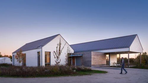 Modern farmhouse-style building with white siding and a metal roof at dusk. A person walks on a dirt path, with trees and a soft pink sky in the background.