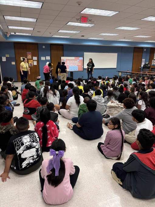 Group of students seated on floor in a classroom, attending a presentation with a projector.