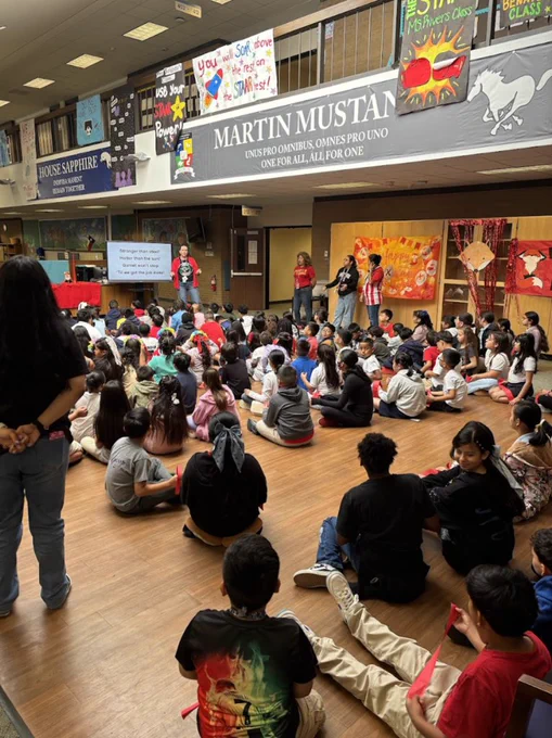 A hall with children sitting on the floor. A banner reads "Martin Mustan". People stand behind a podium.