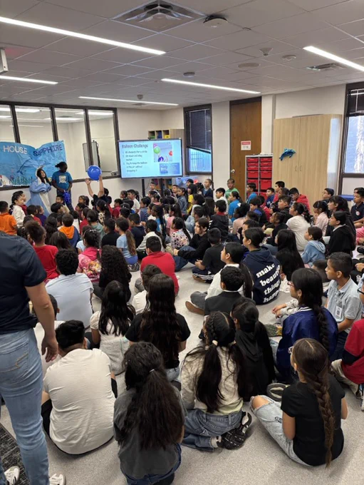 A classroom with students and adults seated on the floor, facing a projector screen on the wall.