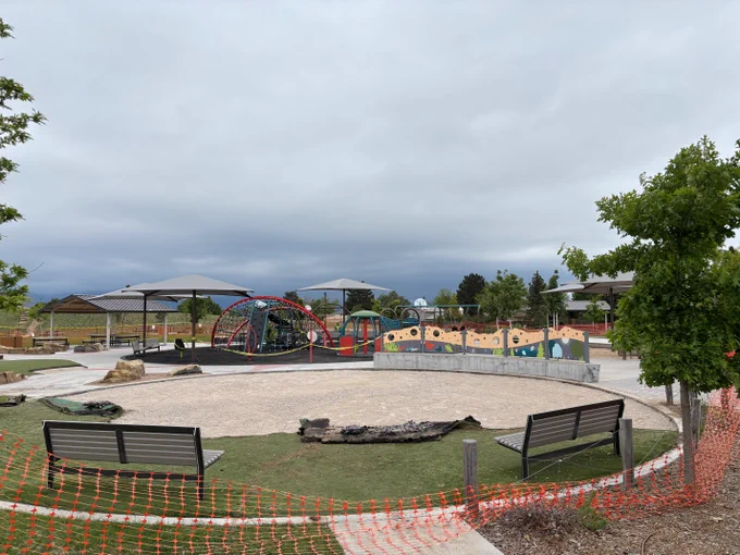 A playground with climbing equipment, slides, and swings is visible under a cloudy sky. There are two shade structures and several benches lining the play area. Red safety netting is on the right side, and trees are scattered throughout the park.