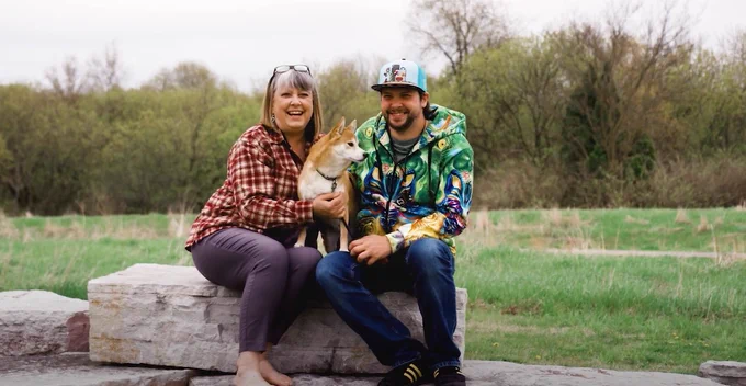 Mother and son sitting on stone bench outdoors holding a dog with trees and grass in the background.
