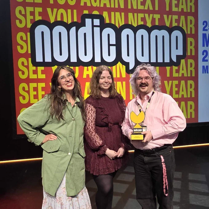 Three smiling people stand in front of a large banner that reads "Nordic Game." One of the people holds a yellow trophy in the shape of a pac-man head.