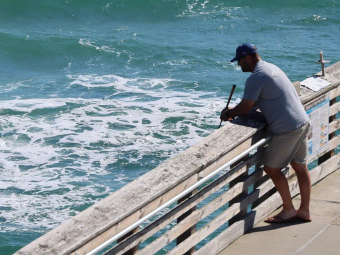 Angler stands on pier overlooking the ocean, engaging in fishing. Waves crash below, and the person leans on the railing, with a fishing rod in hand. The scene is bathed in sunlight.