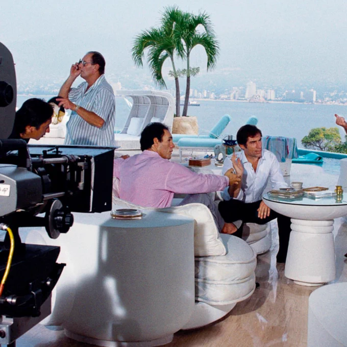 A behind the scenes image from the film Licence To Kill. Timothy Dalton (James Bond) and Robert Davi (Franz Sanchez) are sitting on a balcony with a palm tree in the background. A film camera is visible behind Robert Davi.
