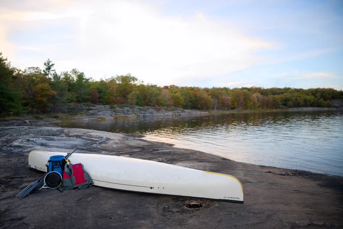 A canoe and gear are placed on a rocky shore, overlooking a calm lake surrounded by trees with autumn foliage under a partly cloudy sky.