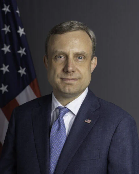 Official portrait of Eric Meyer in a blue suit and patterned blue tie, standing in front of a blurred U.S. flag and gray background, facing the camera with a neutral expression.