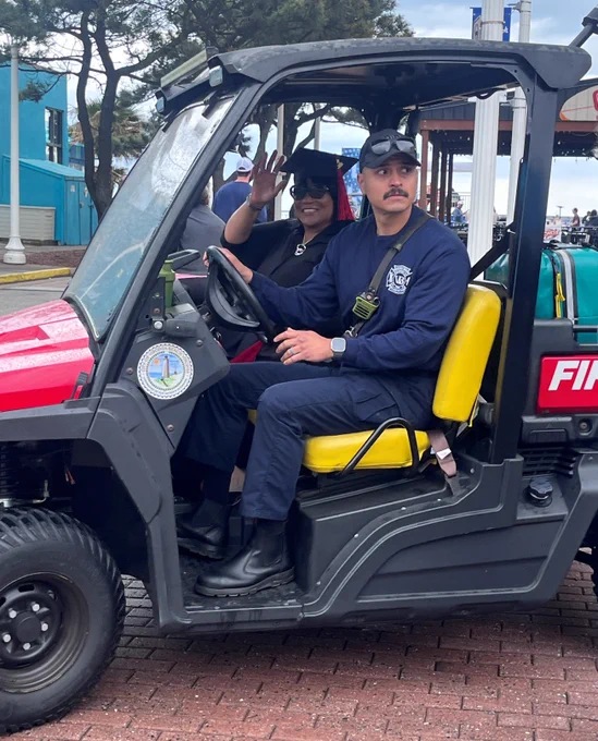 A person in a blue uniform, seated and driving a small emergency services vehicle marked with "Fire." A person in sunglasses is seated next to them, waving. The background features trees, people, and a blue building.