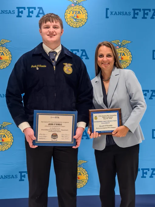FFA student, Jacob O’Trimble, standing on the left hold proficiency award, wearing FFA official dress. Kansas Corn employee, Sharon Thielen, standing on the right holding an award. FFA emblems on the backdrop behind them.