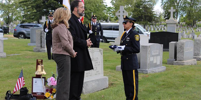 Two people are being presented with a flag at a gravesite. The person presenting the flag is wearing a law enforcement uniform. Two more uniformed officers are standing in the background standing at the position of attention while carrying rifles.