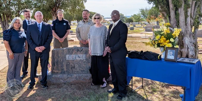A group of seven individuals standing around a grave marker with the name Cline inscribed on it. Two individuals are in law enforcement uniforms. A table with a blue cloth has photos and a bouquet of flowers on it is next to the group.