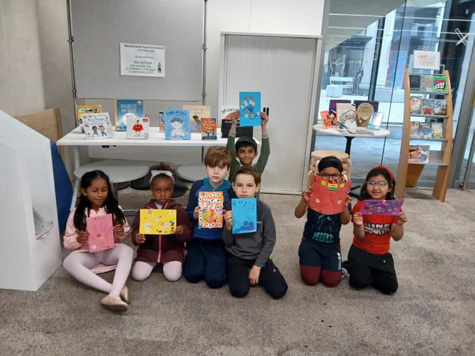 Children joyfully hold up books in front of a colourful table filled with various titles.