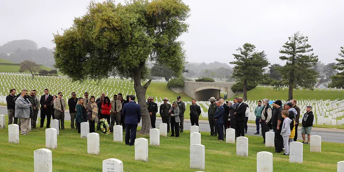A group of people are standing around the headstones inside a cemetery. A wreath of flowers can be seen on the ground next to one of the headstones.