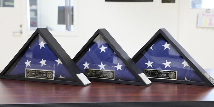 Three folded US flags incased in a triangle picture box frame are positioned on top of a wooden desk. 