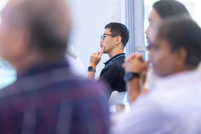A man sits in a large, modern lecture hall and listens as a fellow scientist gives a presentation on his research.