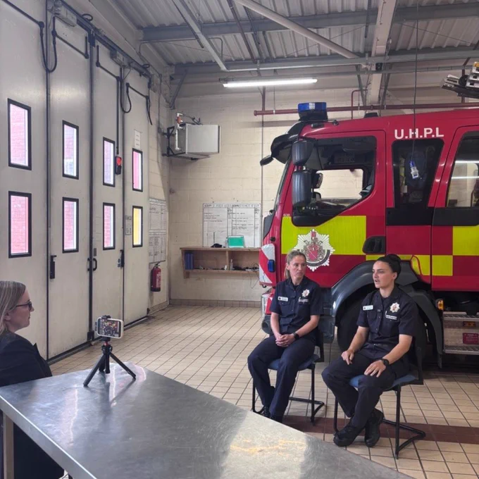 Women sat at table that has on it a camera. Two firefighters in line of the camera with a fire engine in the background