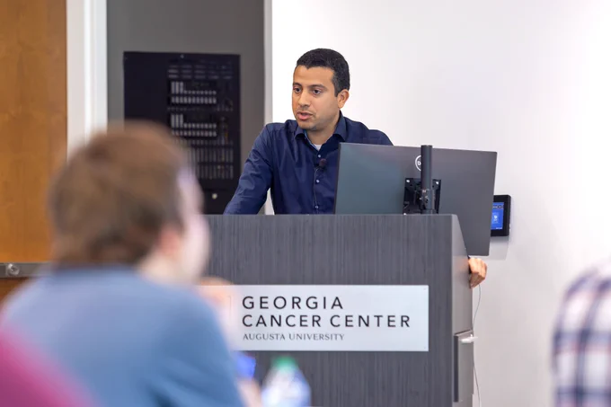 A man stands behind a podium with a computer screen and gives a presentation on his scientific research to a room full of fellow scientists and trainees.