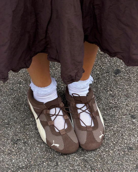 An image of a person's lower legs wearing a pair of brown PUMA Speedcat Go trainers with a brown skirt whilst standing on a concrete ground.