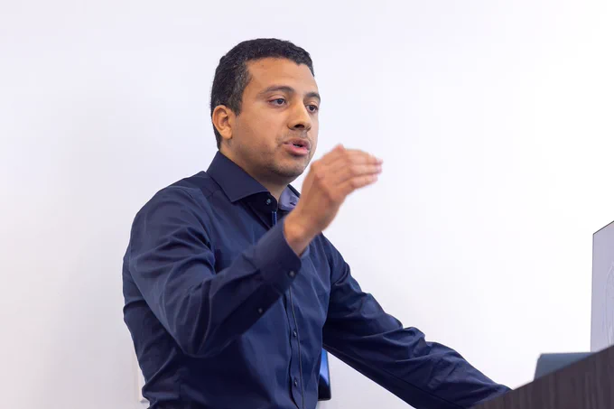 A man stands behind a podium with a computer screen and gives a presentation on his scientific research to a room full of fellow scientists and trainees.