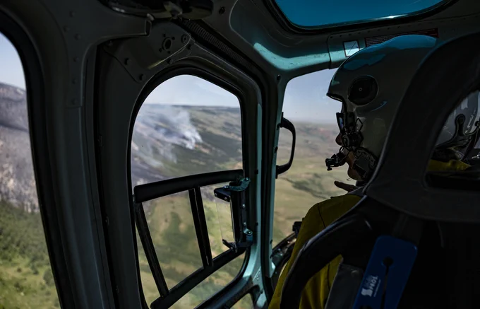 View from inside a helicopter cockpit showing a pilot wearing a helmet flying over a landscape with smoke rising from a distant wildfire.