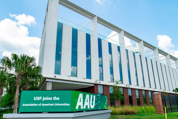 Modern university building with tall vertical windows and a green sign announcing USF joining the Association of American Universities.