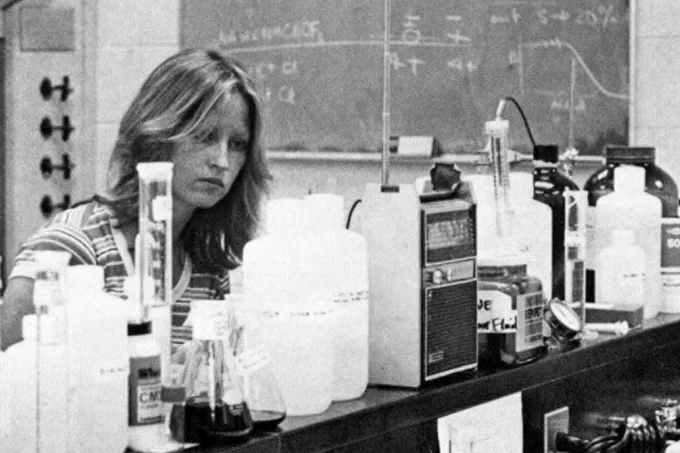 A scientist working in a laboratory surrounded by various chemical bottles, flasks, and scientific equipment on a cluttered bench.