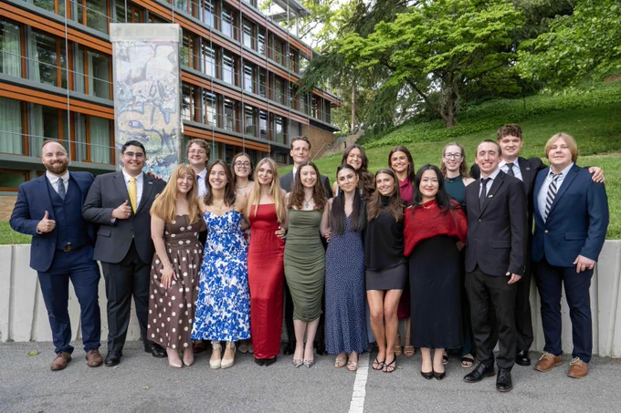 A group of people stands together outside a modern building, smiling at the camera. They are dressed in formal attire, with a mix of suits and dresses. The background features a building with large windows, greenery, and a painted pillar.