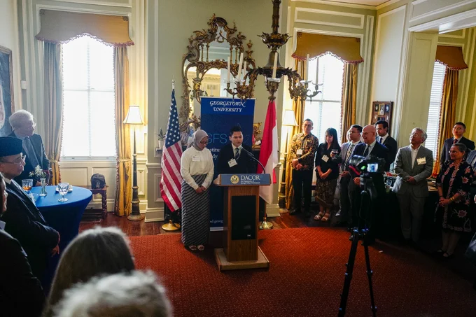 A person is speaking at a podium in a formal room, with a "Georgetown University" banner in the background. An audience stands around, including people in business and formal attire. American and another flag are visible nearby. The event is being recorded with a camera on a tripod.