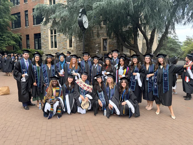 Graduates in caps and gowns pose in front of a historic building with a clock, smiling and holding diplomas. Elegant trees and brick pathways surround the scene.