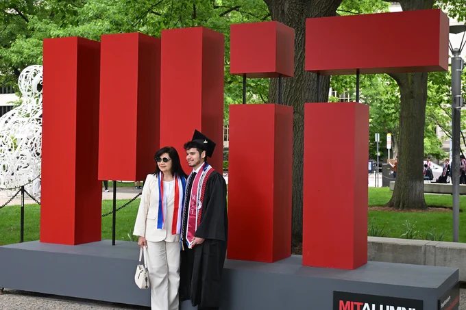 An MIT graduate stands in front of a large MIT logo.