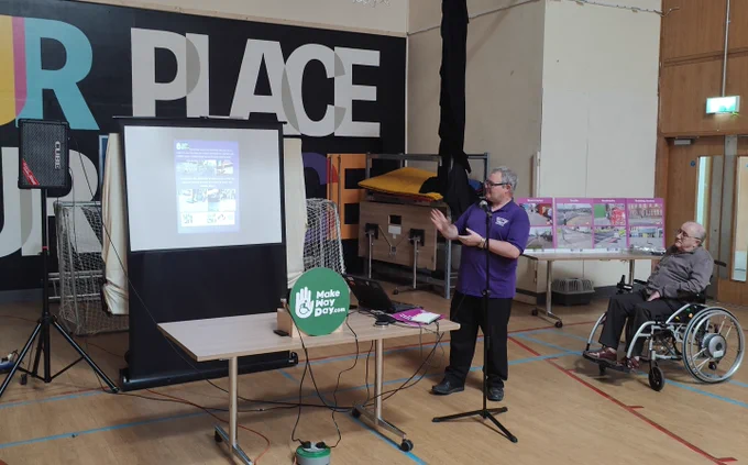 A speaker in a purple polo shirt addresses attendees at a Make Way Day event in a sports hall, with a projected presentation visible on a screen behind him. A green Make Way Day sign sits on a table in the foreground. To his right, a man using a powered wheelchair listens to the presentation. A display board with images is visible in the background, and a large mural reading "Our Place" covers the