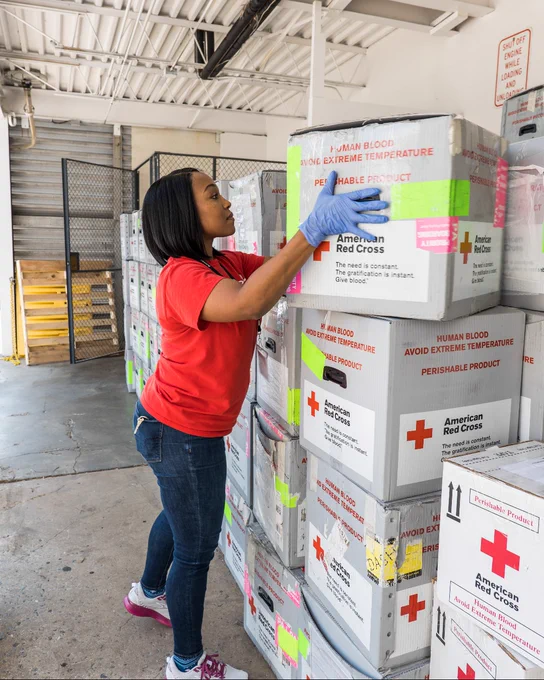 A person in a red shirt stacks boxes labeled "American Red Cross" and "Human Blood" in a warehouse area. The boxes have warnings about temperature and perishable contents. The environment includes a loading dock.