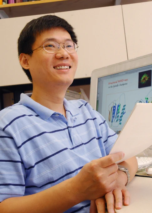 Dr. Hongtao Yu in his lab. He is wearing a blue and white striped polo and is holding papers. The screen behind him shows 3D animations of his research.