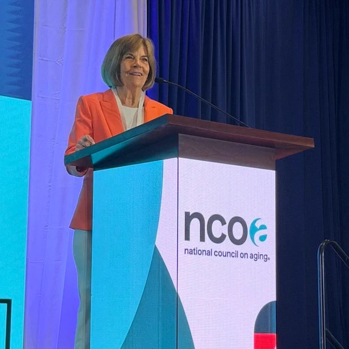 Mary Lazare, smiling in a tangerine blazer, stands at an NCOA-branded podium in front of draped background