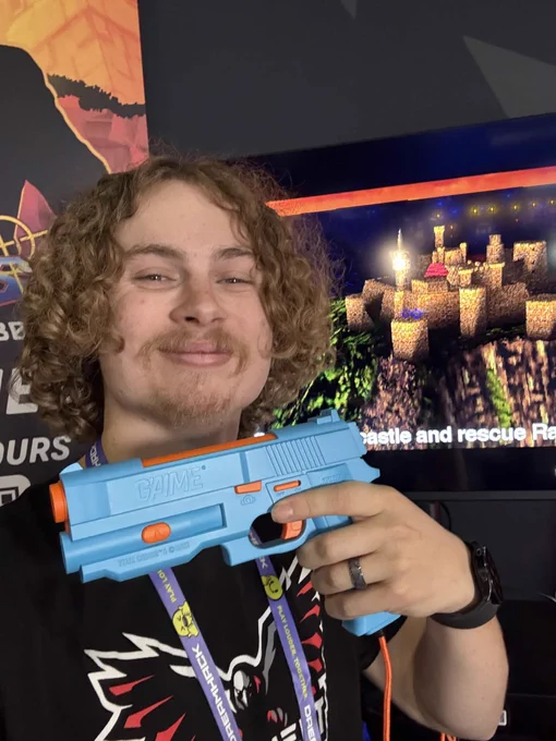 A person holds a blue toy gun while smiling at a gaming event. A pixel art castle is visible on the screen in the background.
