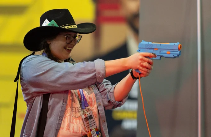 A woman wearing a cowboy hat and glasses enjoys herself while aiming a blue and orange toy gun at a gaming event. Her excitement is evident in her smile.