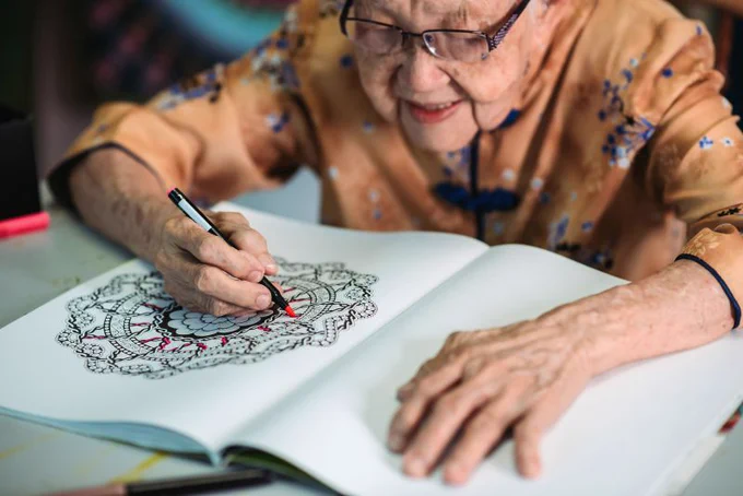 An elderly woman colors as intricate design in a coloring book.