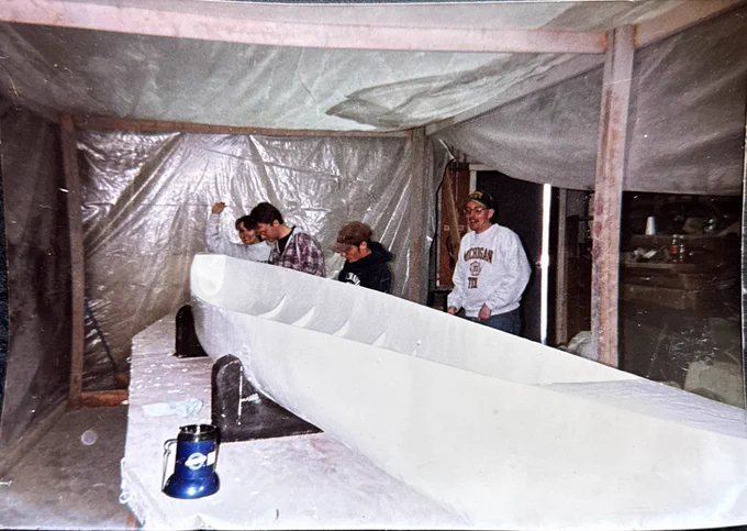 Several Michigan Tech Concrete Canoe team members stand around a large canoe mold inside a workshop enclosed with plastic sheeting. The canoe is in an early stage of construction, and the team appears to be inspecting or shaping the mold.