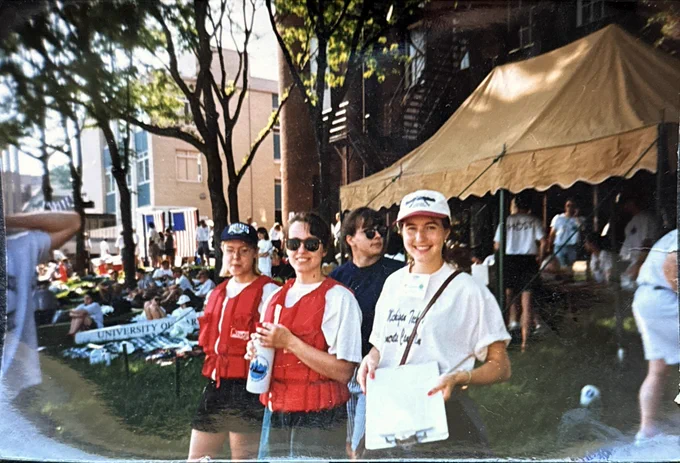 A group of Michigan Tech Concrete Canoe team members stand outdoors at a competition venue wearing life jackets and team apparel. Tents, spectators, and other canoe teams are visible in the background.