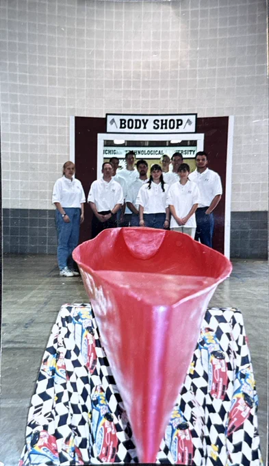 A red concrete canoe sits on display in the foreground inside a large arena. Behind it, a group of Michigan Tech team members pose together in matching white polo shirts in front of a display labeled "Body Shop."